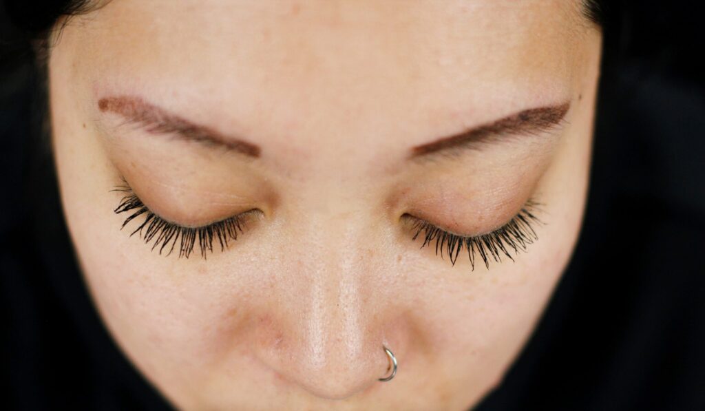 Close-up of a person's closed eyes with long eyelashes and a nose piercing.