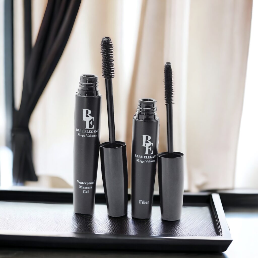 Two black mascara tubes with wands on a black surface near curtains.